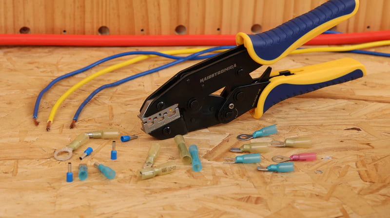 Gloved technician crimping wire connector using Haisstronica tool beside organizer box filled with assorted terminals
